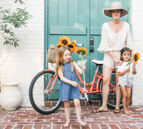 Our super soft, lightweight double gauze Pocket Tee in the color Shell paired with Racer shorts in Bloom. Another girl is wearing our Crossback Ruffle dress in Ocean while holding a sunflower. Their beautiful mother is wearing a swimsuit cover up and a hat.