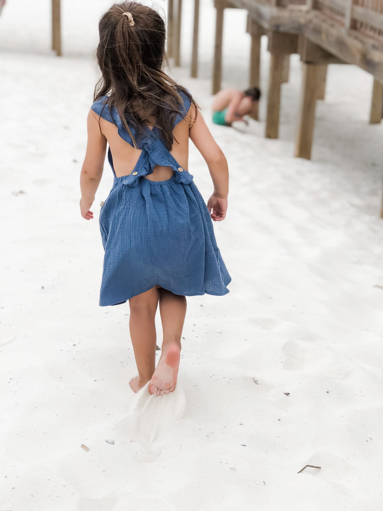 Girl wearing our signature double gauze cross back ruffle dress in the color Ocean while playing in the sand.
