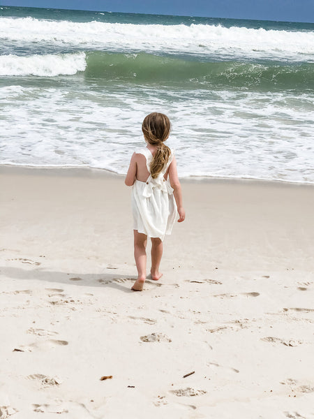 Girl watching the waves standing in the sand while wearing our signature double gauze cross back ruffle dress in the color shell.